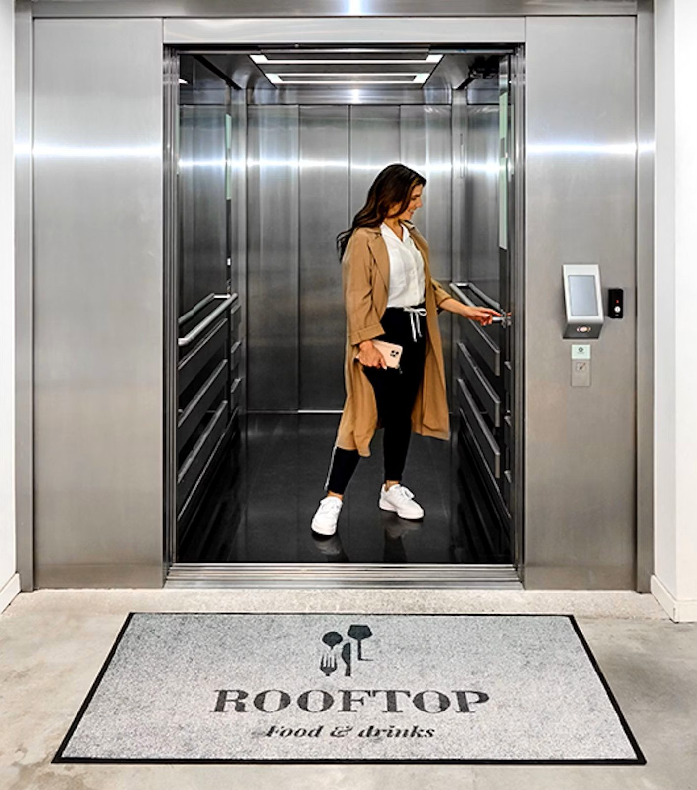 Frau steht im Aufzug. Vor dem Aufzug liegt eine Logo-Matte mit der Aufschrift Rooftop.